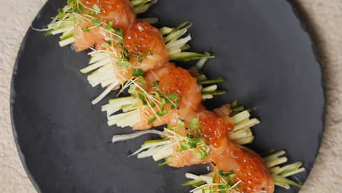 Five salmon rolls, topped with orange caviar and microgreens, in a row on black dish