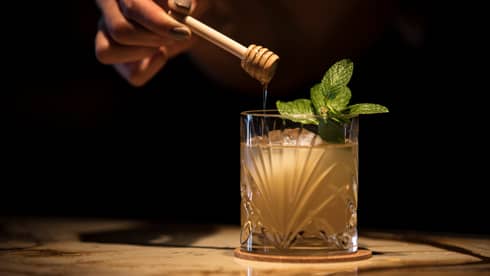 A cocktail with a sprig of mint sits on a table with dim lighting, a hand drizzles honey into it