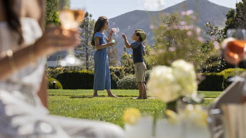 Two children blowing bubbles outside in the distance while a woman drinks a glass of wine in the foreground.