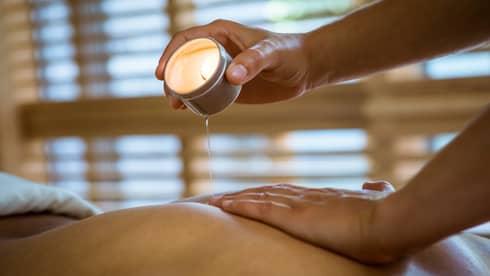 Massage therapist pours hot wax from tealight candle over person's bare back in spa