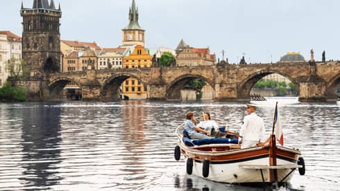 Couple with captain in wooden boat on Vltava River with bridge and buildings in backdrop