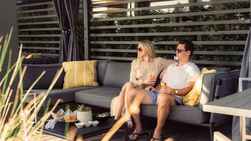 Two people sit on a patio couch with drinks, surrounded by cushions and snacks on a table. They wear sunglasses and casual summer attire.