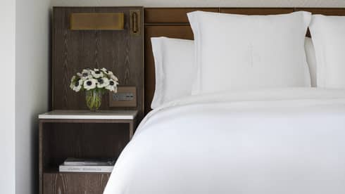 White bed with Four Seasons-embroidered pillows, set next to a wooden night stand with flowers