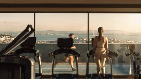 A person uses a treadmill facing floor-to-ceiling windows in a rooftop fitness centre. Another person is running by on the track outside, and the coastline and water are visible in the background.
