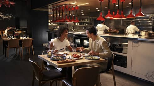 Two people sit at a square dining table in front of an open kitchen with two chefs working behind them