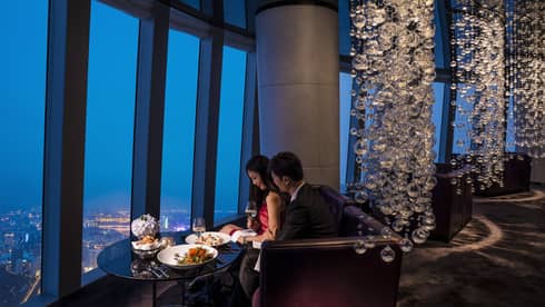 Man and woman with wine dine at romantic cocktail table by window looking out at city lights