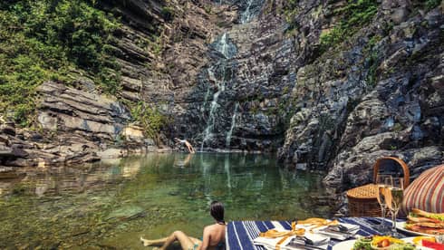 Woman wades in water at bottom of waterfall by picnic lunch set up on rocks