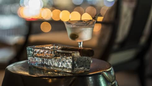 Cigar in heavy rectangular glass ashtray beside martini glass with olive 