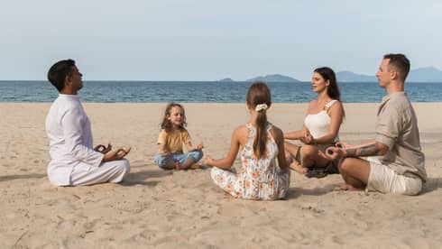 A family meditating together on a sandy beach, guided by an instructor, with the ocean and distant islands in the background.