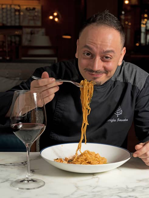A man in a black Four Seasons shirt with a forkful of spaghetti noodles and a glass of wine.