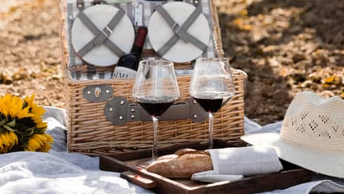 An open wicker picnic basket, two glasses of red wine and a baguette sit on a white blanket among rows of glowing vines.