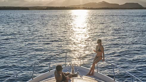 A couple in swimwear relaxing on the bow of a boat, wine glasses in hand and the setting sun glinting off the endless ocean.