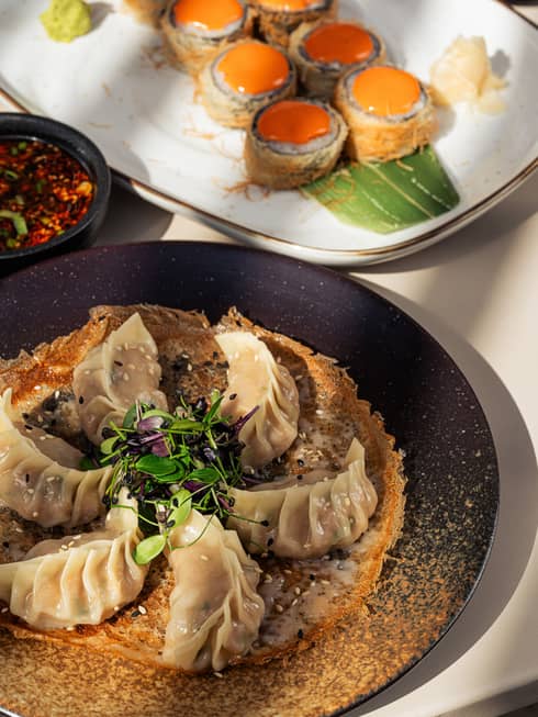 A blac, round plate filled with six dumplings and garnish sits next to a roll of sushi plated on a white rectangular dish