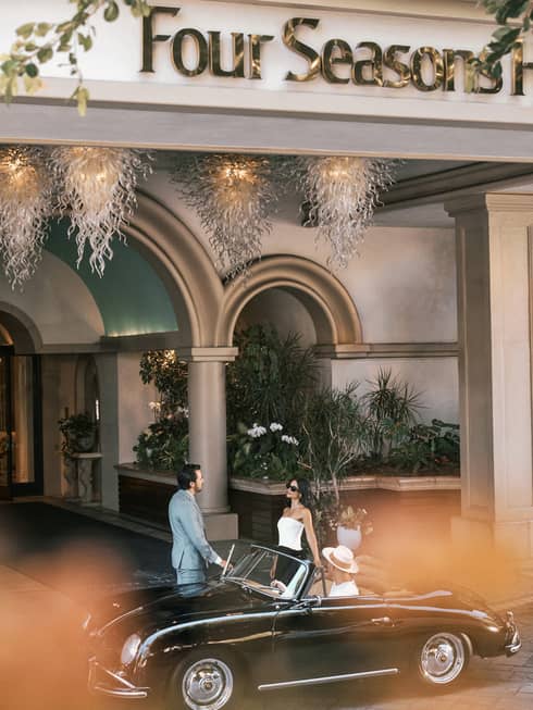 A valet helps a couple into their black sports car in front of a hotel entrance