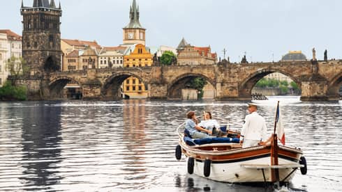 Couple with captain in wooden boat on Vltava River with bridge and buildings in backdrop