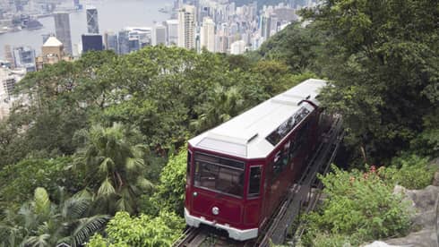 The red Peak Tram ascends a lush, green hillside with a sprawling cityscape in the background. Tall skyscrapers rise into the skyline under a cloudy sky.