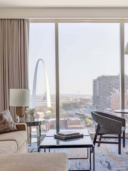 Luxury hotel room with a beige sofa, coffee tables, chairs, and floor-to-ceiling windows with a view of the Gateway Arch and St. Louis city buildings
