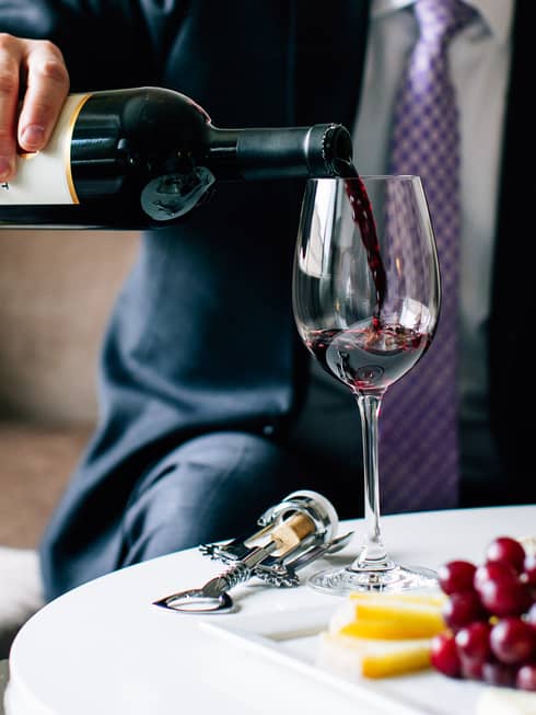 Close-up view of poured red wine cascading into a glass, held by a person with a jacket and tie.