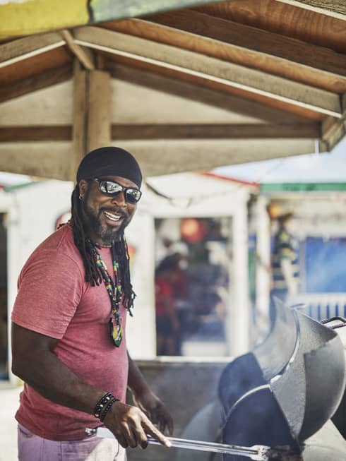 Side view of a smiling colourfully dressed chef barbecuing meat under a wooden canopy, the air hazy with sunlight and smoke. 