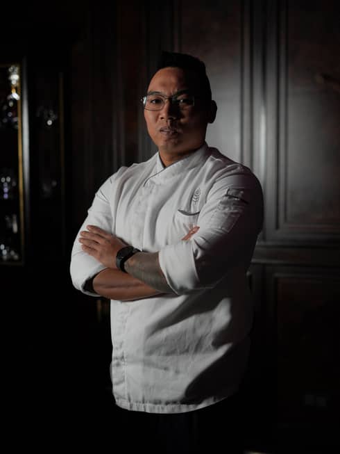 A chef wearing a white shirt stands with arms crossed in an interior space