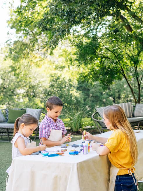 Two children and a young woman are painting rocks outside on a table, large trees surround them.