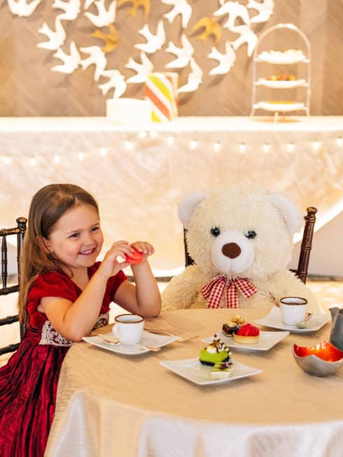 A small girl in a red dress sitting with a large teddy bear at a table with tea and small desserts.