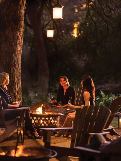 Three women sitting around a fire drinking wine outside.