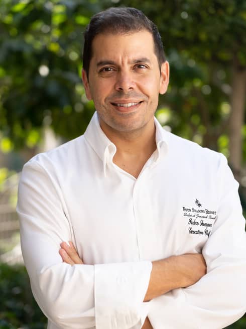 A chef in a Four Seasons uniform with his arms crossed in front of a canopy of foliage.