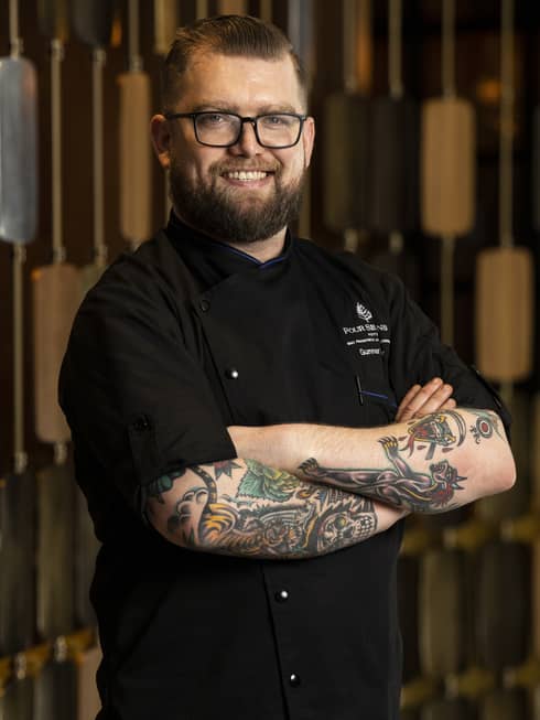 A chef in a black chef's coat with tattoos on his arm.