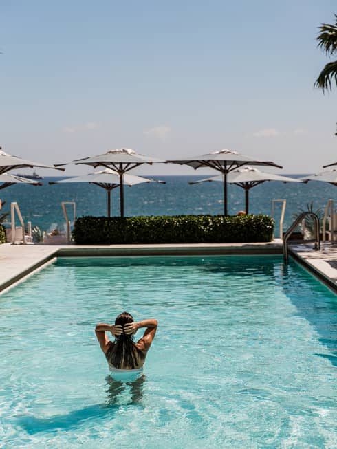 Person stands in a rooftop pool with two rows of umbrellas nearby and a scenic view in the distance