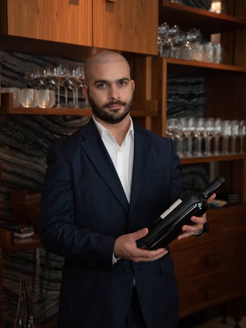 Sommelier David Lopes in black suit and white shirt, standing and holding a bottle of wine