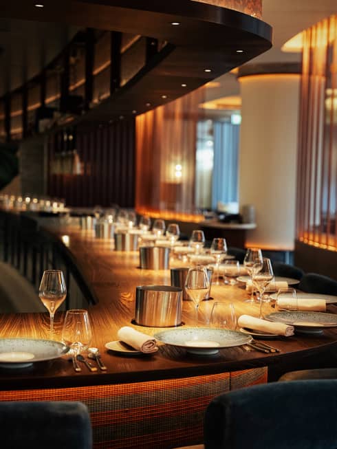 A long, curved solid wood bar with a dozen place settings in warm, golden lighting.