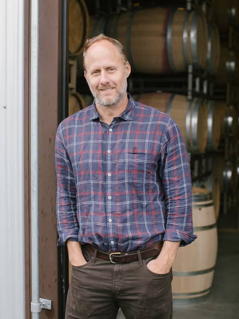 A man in a plaid shirt standing in front of wine barrels.