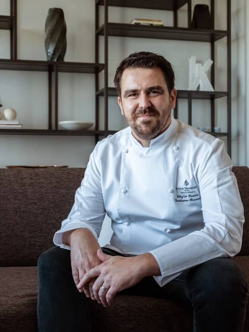 Smiling, bearded man wearing white chef's coat poses for camera while sitting on couch