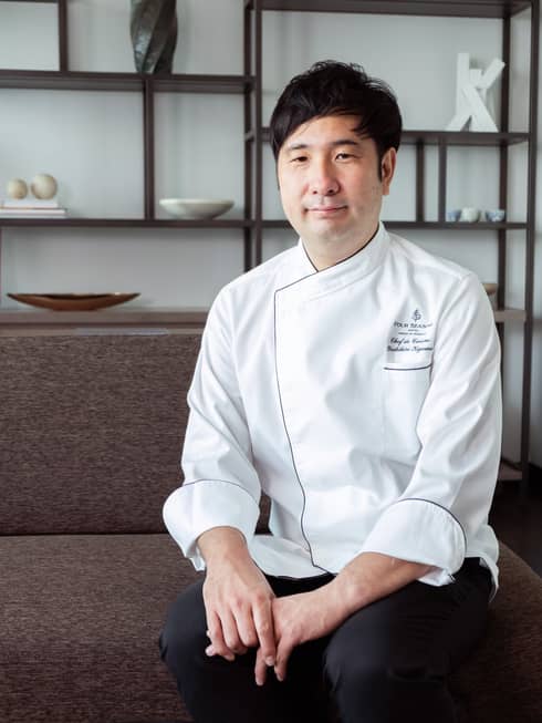 Chef de Cuisine Kigawa, wearing a white chef's coat, smiles for the camera while sitting on a couch