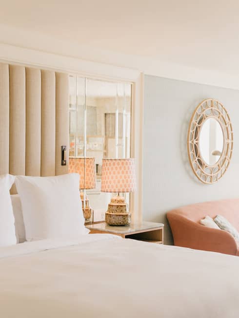 Elegant hotel room with a plush white bed, mirrored headboard, stylish lamps on nightstands, and a round decorative mirror on the wall.