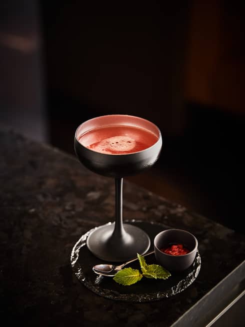 Red cocktail in pewter saucer champagne glass garnished at the base with strawberries and mint leaves