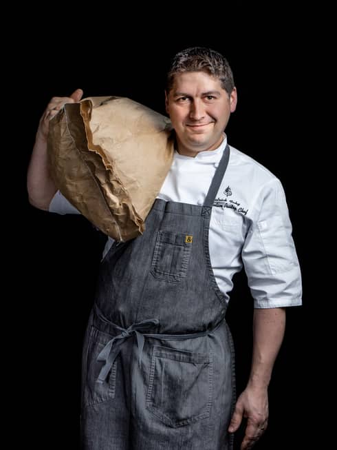A chef in a grey apron holding a large brown bag.