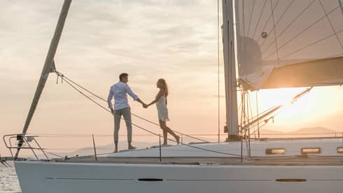 A smiling couple holding hands while they walk across the deck of a sailboat illuminated from behind by the setting sun.