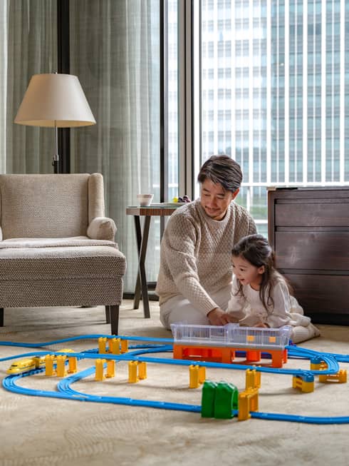 Man and young girl play with toy train set in guest room living area