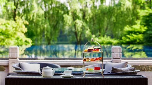 Afternoon tea tray with sweets, teapot, cup on patio table, garden trees in background