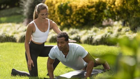 A yoga instructor helping a man get into a yoga position outside on very green grass.