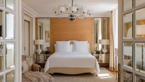 Luxury hotel bedroom with a king bed, mirrored bedside tables with lamps and a modern chandelier, complemented by soft beige accents and an elegant armchair