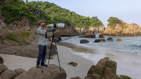Photographer stands behind large camera lens on a rocky beach