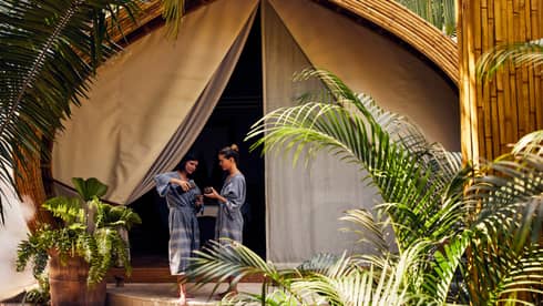 Two women in spa robes stand outside a luxury spa tent, one pouring tea for the other, surrounded by lush greenery in a peaceful setting.