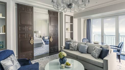 Sea-view living room, with grey sofa, blue pillows and accent chairs, chandelier, dark wooden doors open to bedroom