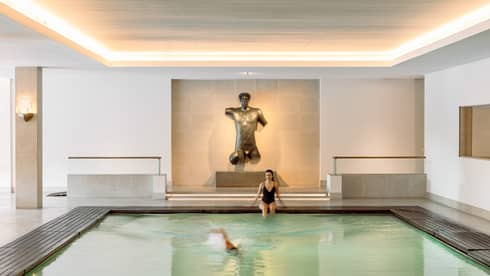 Indoor pool with a swimmer and a person sitting at the edge, featuring a large bronze torso sculpture on the far wall.
