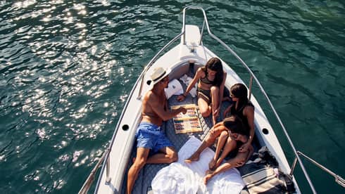 A family lounges in the bow of a luxurious boat amid towels and blankets, playing backgammon, the sun's glow on teal waters. 