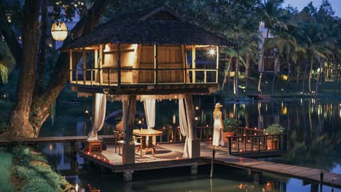 Person standing on a dock beside a candlelit lakeside pavilion with a thatched roof and draped curtains at dusk.