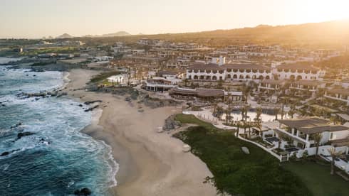 Aerial view of a Cabo San Lucas luxury beachfront resort with white buildings, palm trees and sandy coastline at sunset